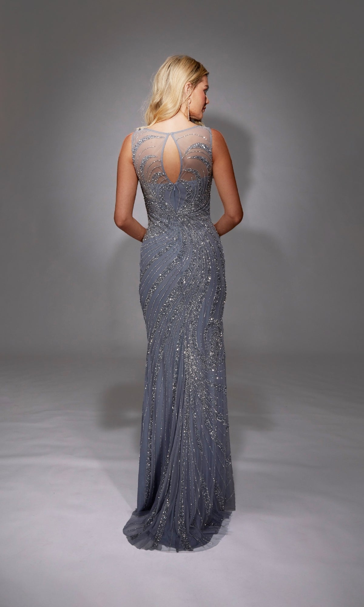 A woman faces away, wearing the Long Formal Dress: Alyce 27846—a floor-length silver-gray sheath with intricate beading and a sheer back panel featuring a keyhole detail.